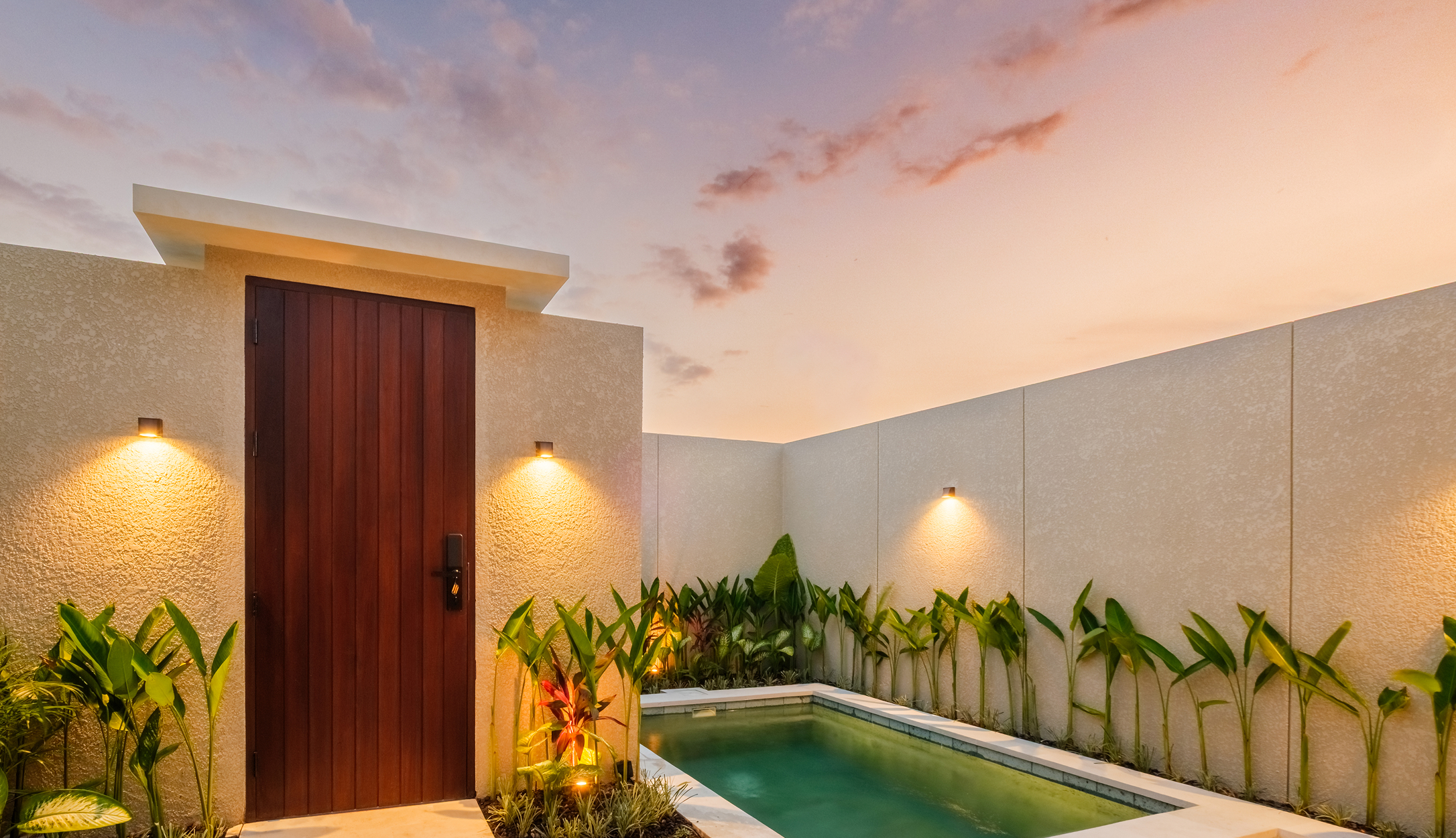 Timber villa entrance door beside the private plunge pool, with wall lights glowing at sunset