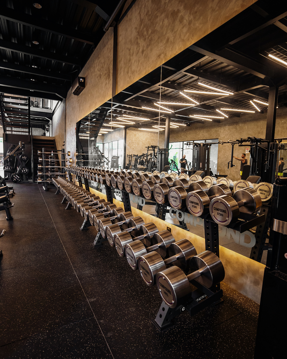 Rows of dumbbells along a mirrored wall in The Compound gym under linear ceiling lights