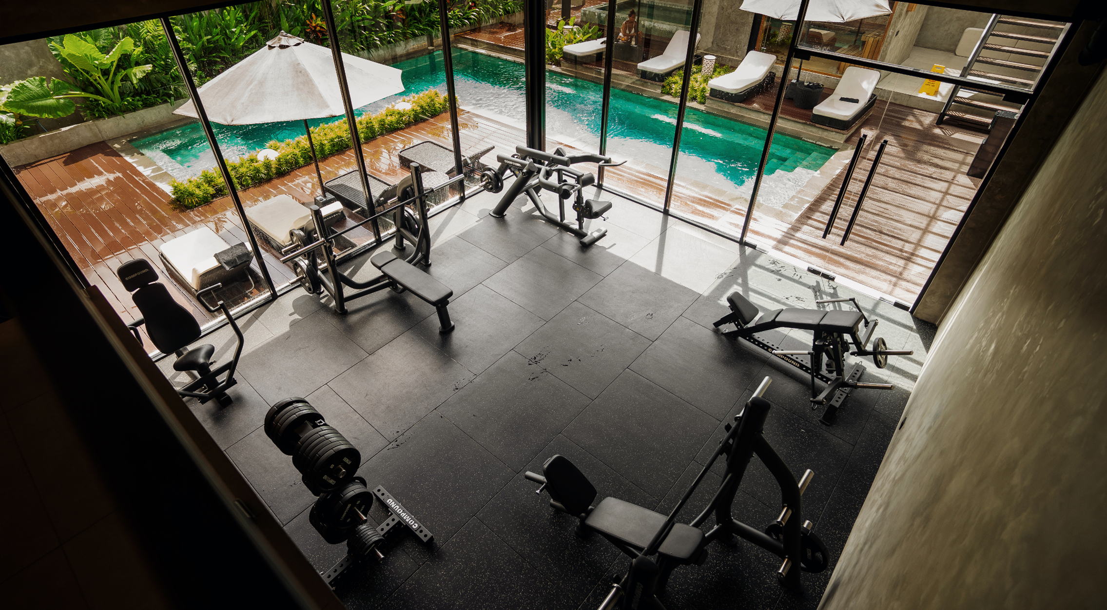 View down into The Compound's glass-walled gym, overlooking the pool, umbrellas and sun loungers